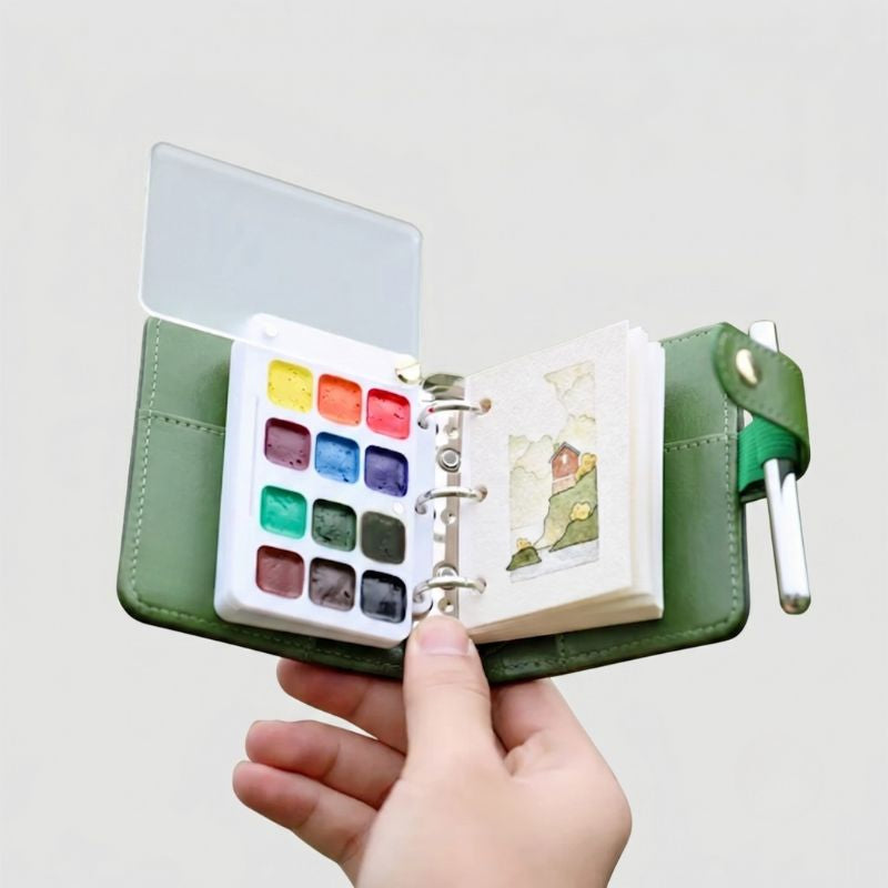 Hand holding the open ArtMate Nomad Mini Kit in green leather, with a 12-pan palette and a small landscape painting