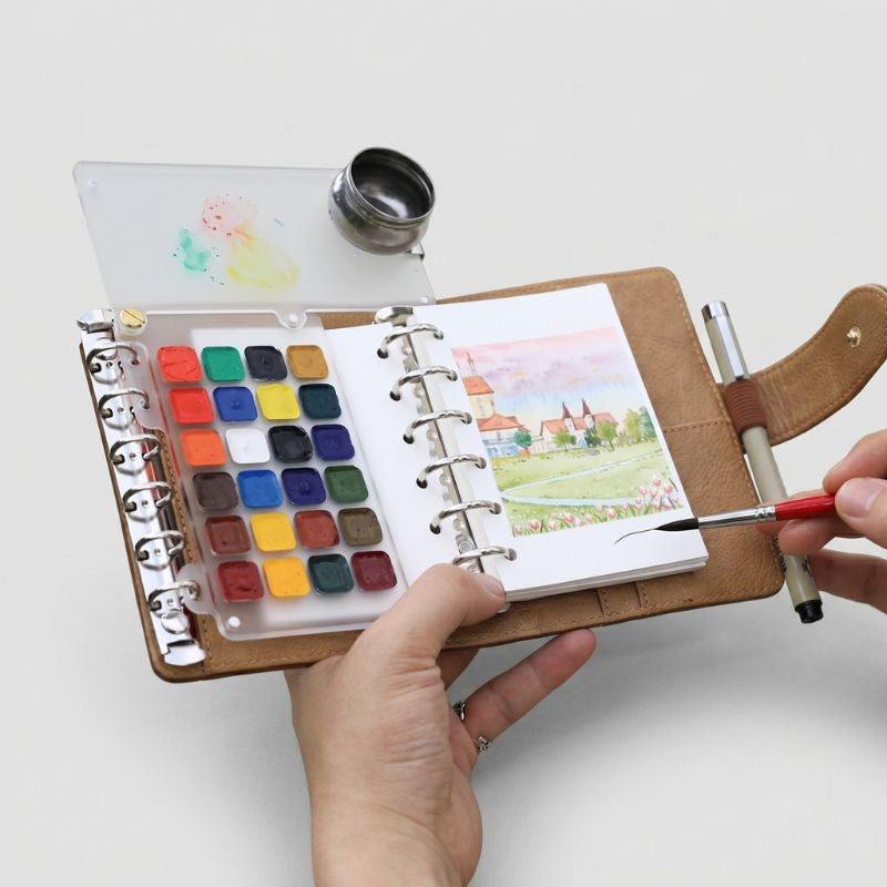 Hands holding the open ArtMate Nomad leather watercolor kit in a meadow, 24-pan palette and village painting